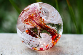 white and red glimmer orb with cremation ash sitting on stone in front of bush